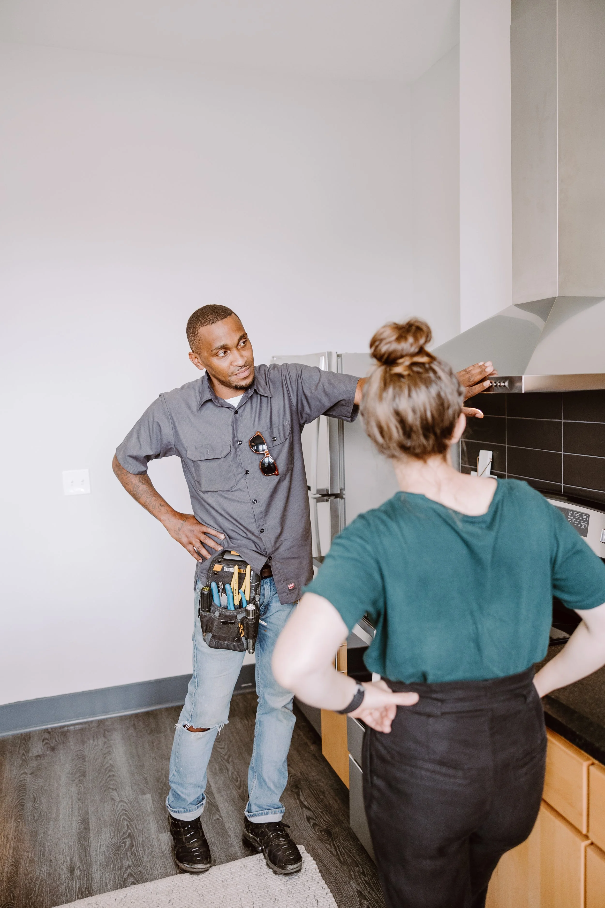 A man with a tool belt talking to a woman in a kitchen, both with hands on the counter, engaging in a discussion about kitchen repairs.
