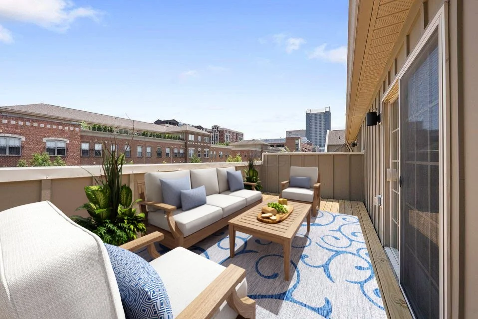 Outdoor balcony with sofa, armchair, and coffee table, blue patterned outdoor rug, potted plants, and cityscape view during daytime.