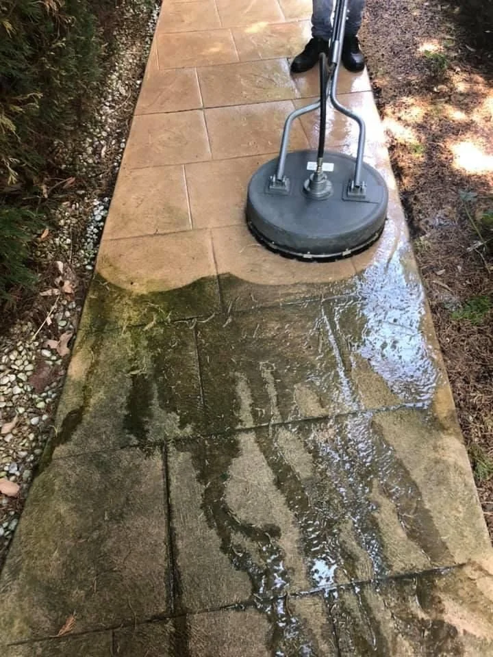 A pressure washer cleaning a stone sidewalk, with water and soap being sprayed on the surface. A person’s legs and black shoes are visible in the background.