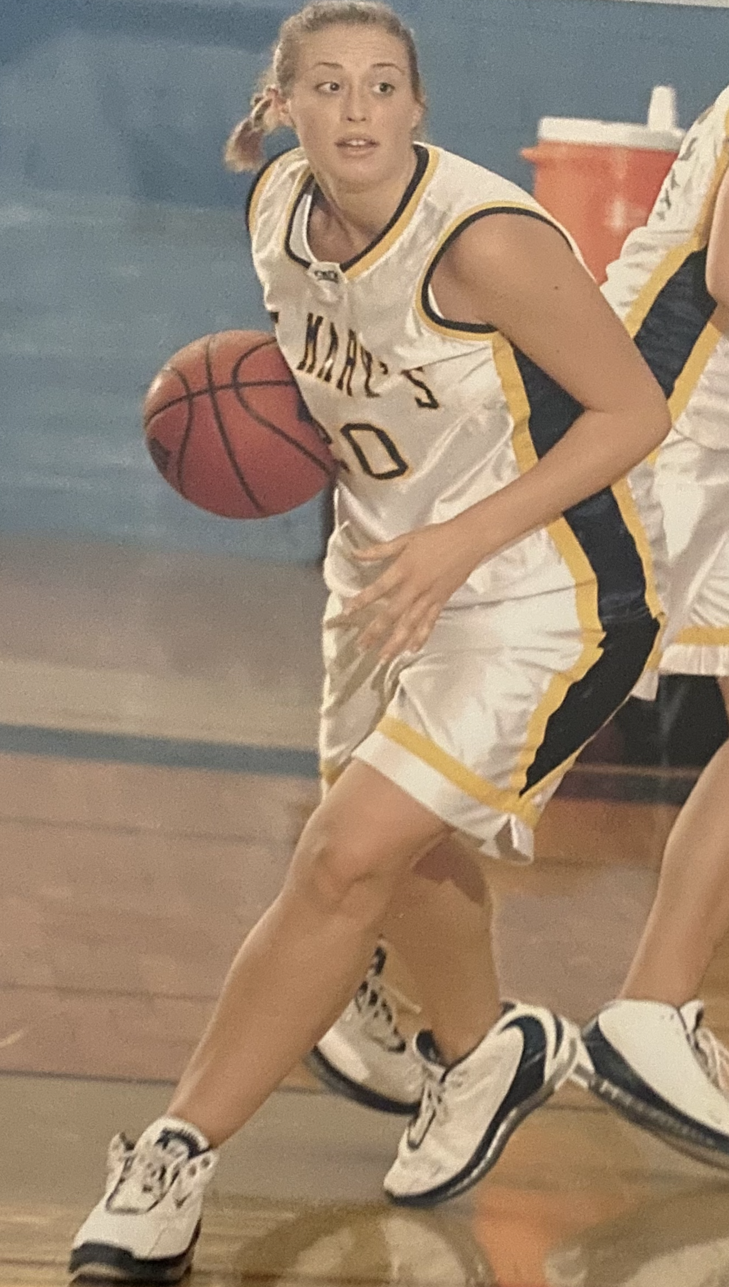 A young woman playing basketball on an indoor court, wearing a white sports jersey and shorts with black and yellow accents, holding a basketball, and wearing white basketball shoes with black details.