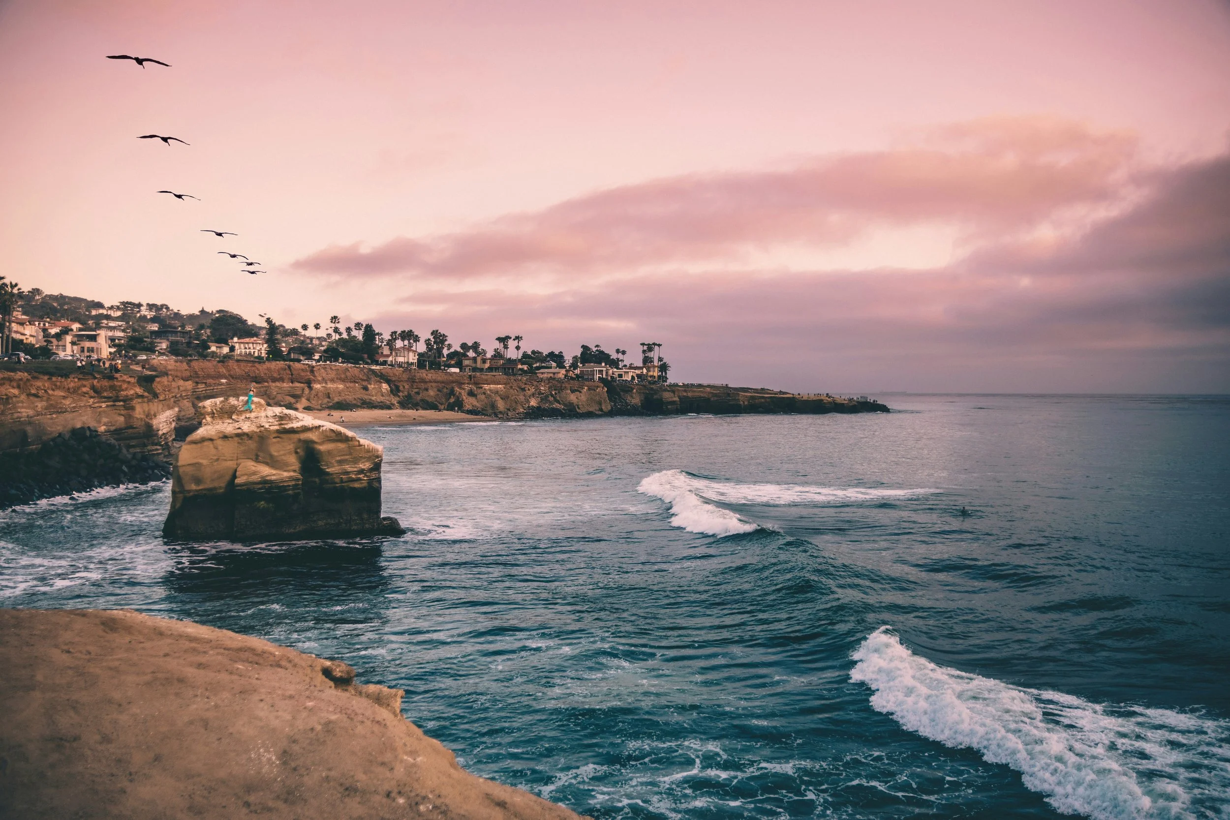 Scenic view of the ocean with crashing waves, a large rock formation near the shore, distant houses along a cliff, and seagulls flying in a partly cloudy sky during sunset or sunrise.