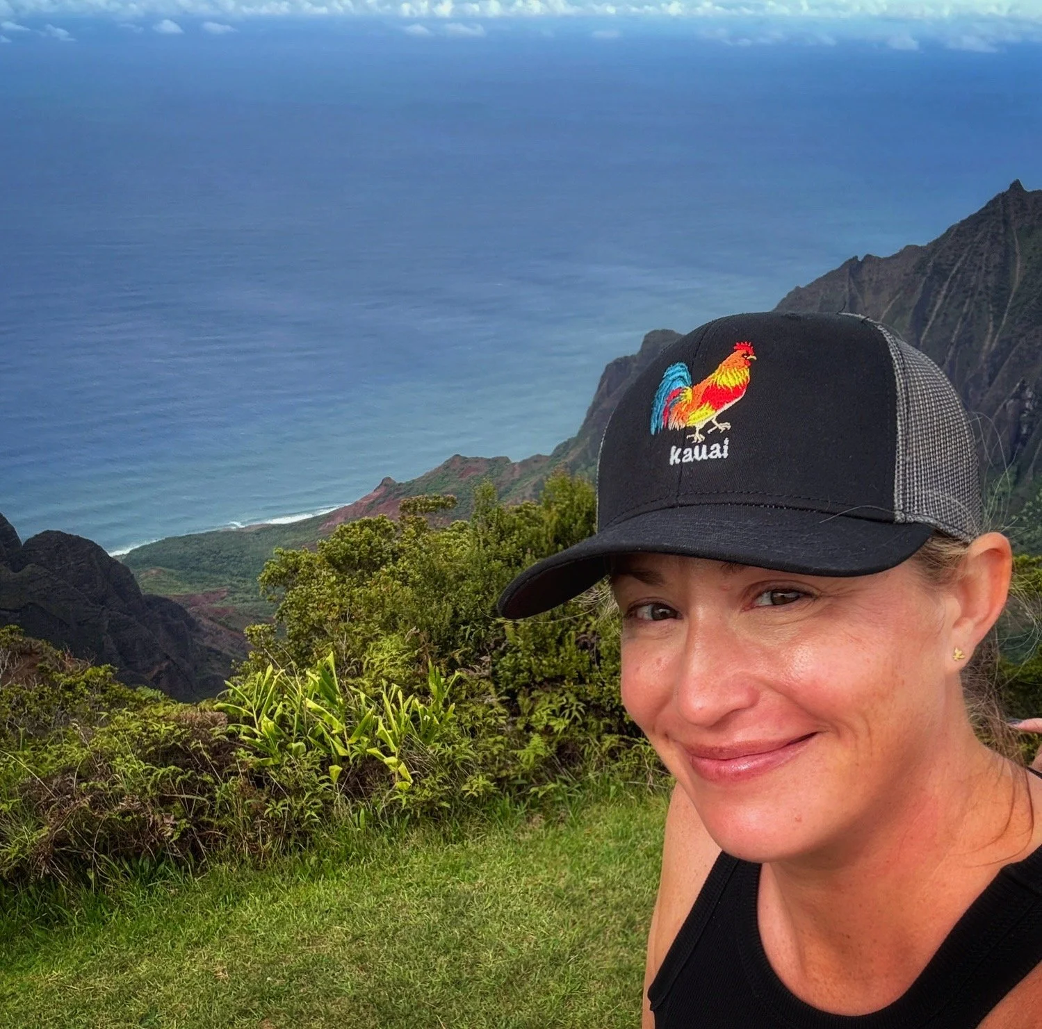 A woman smiling in front of a coastal landscape with green hills and the ocean, wearing a black cap with a colorful rooster and the word 'Kauai' embroidered on it.