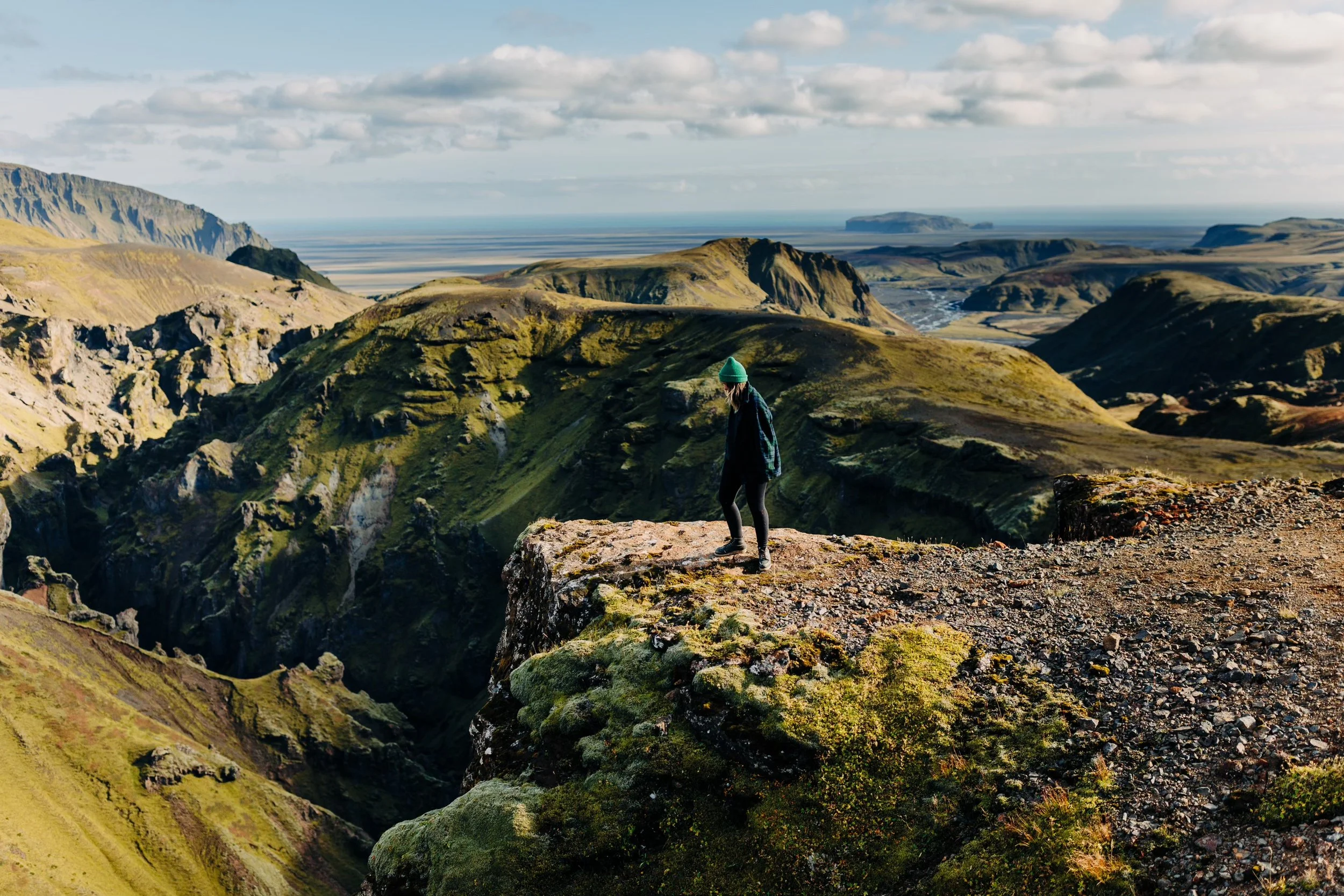 Person standing on rocky edge overlooking volcanic landscape with green moss, hills, and distant ocean under cloudy sky.