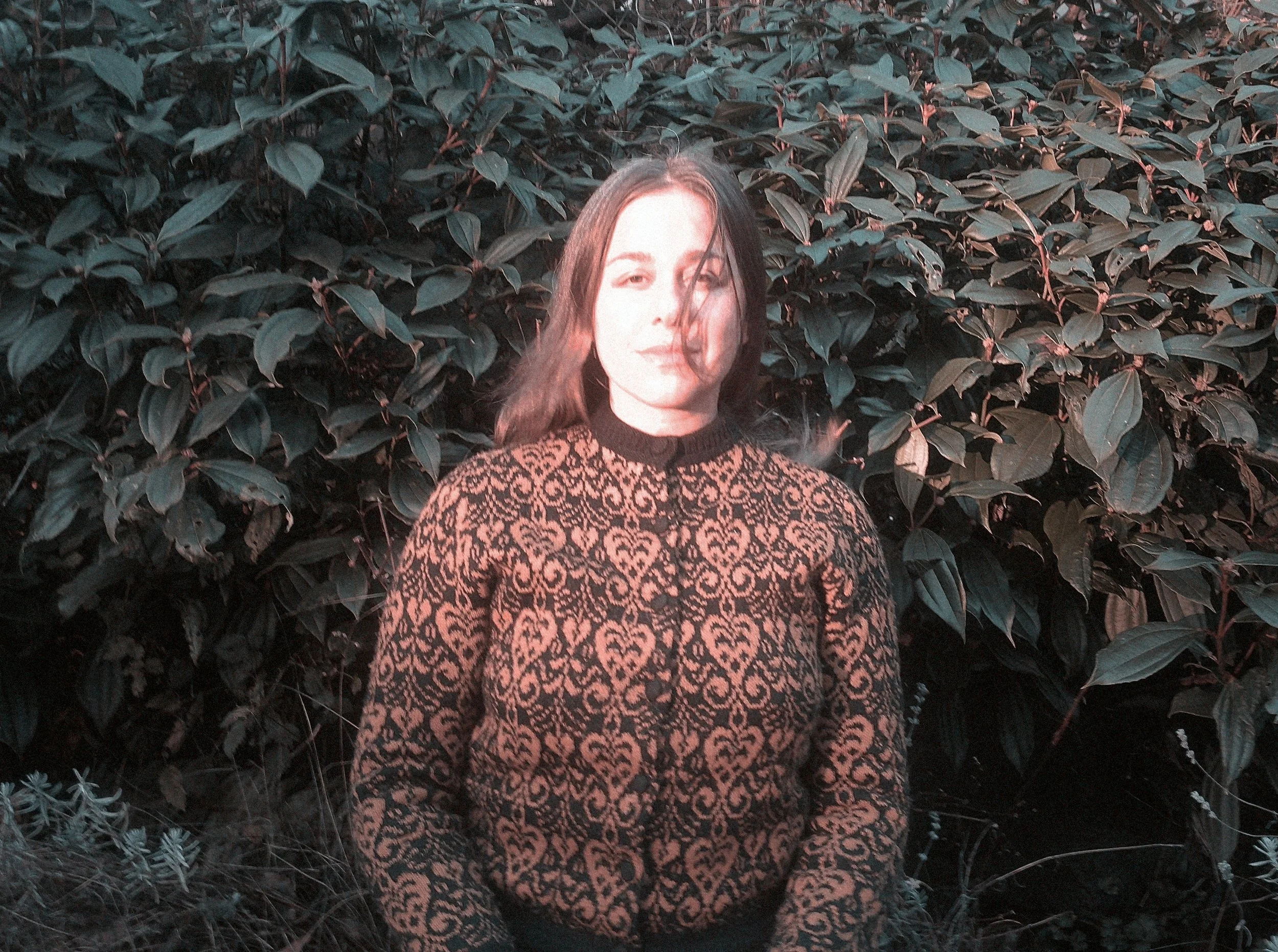 A woman with shoulder-length hair stands in front of dense green foliage, wearing a black and brown patterned sweater.