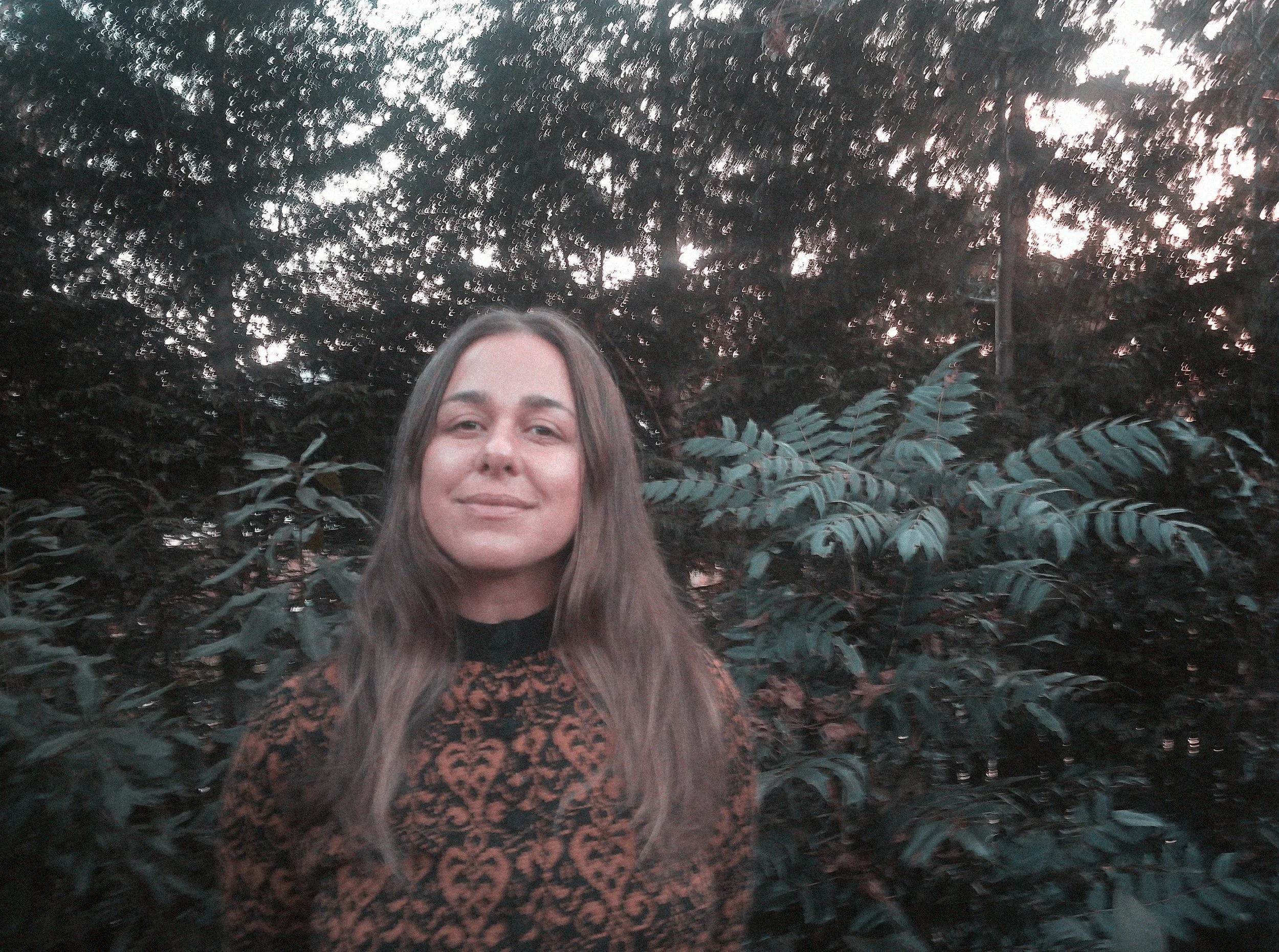 A young woman with long brown hair standing outdoors in front of dense dark green foliage, with the sky visible through the trees in the background.