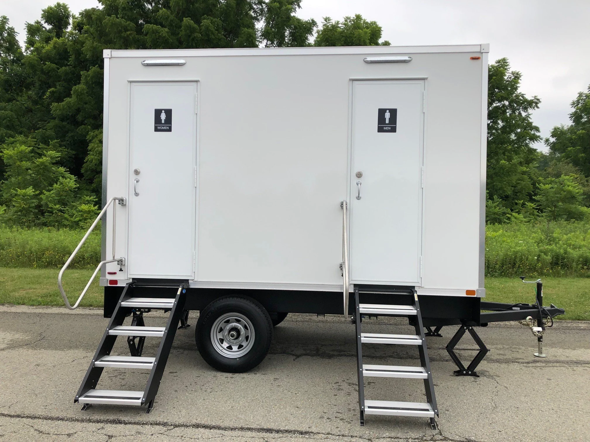 White portable restroom trailer with separate entrances for women and men, each with portable stairs and handrails, situated on a paved surface with green trees in the background.