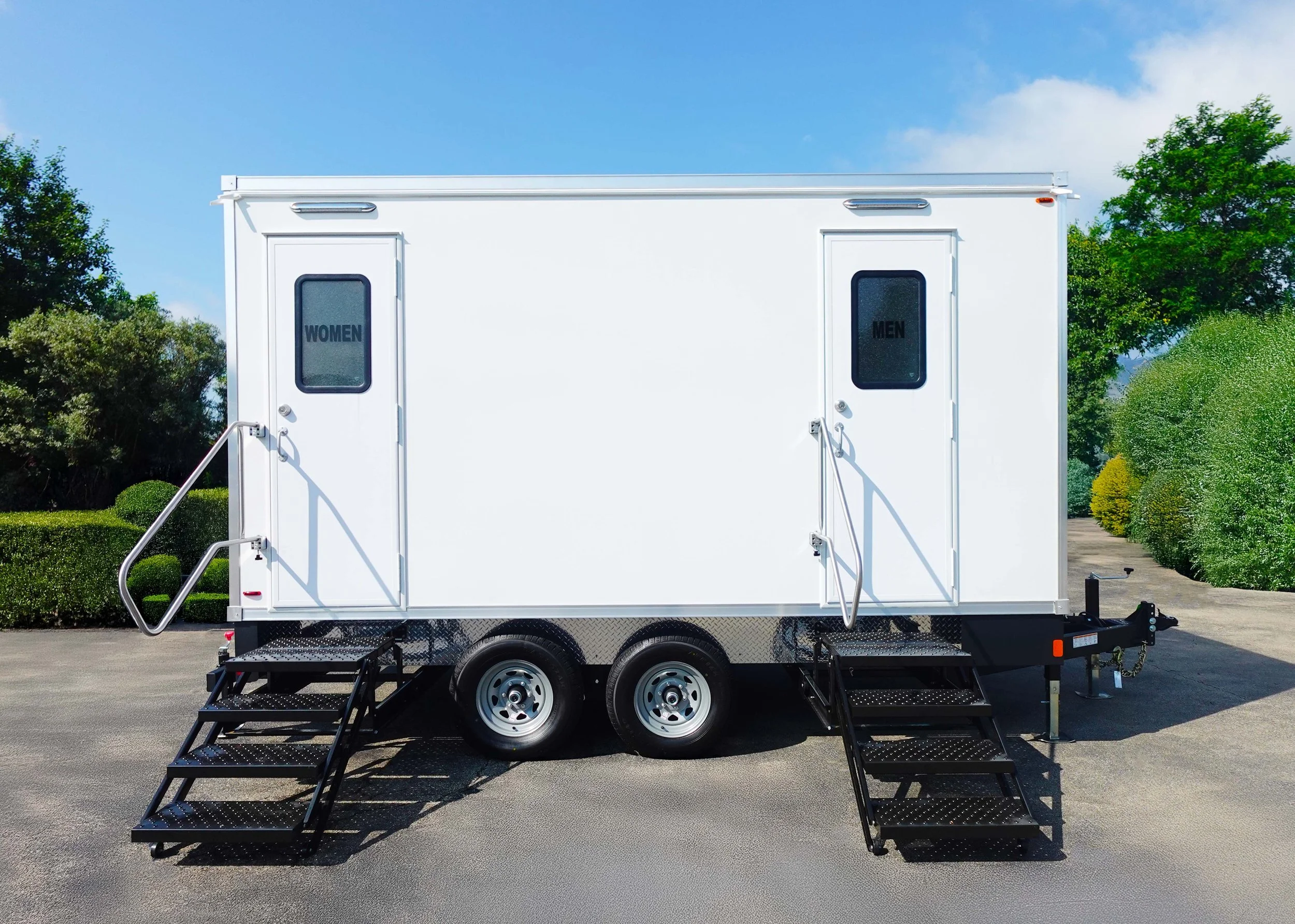 A white mobile restroom trailer with two separate doors labeled WOMEN and MEN, four stairs leading up to each entrance, and surrounded by greenery under a partly cloudy sky.