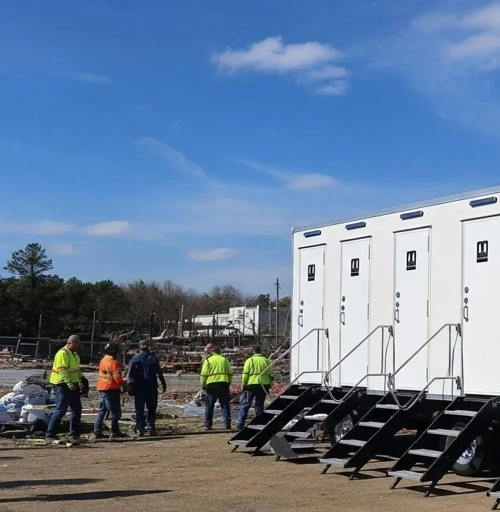 A row of portable restroom trailers on a dirt lot with a group of workers walking nearby, under a blue sky with some clouds.