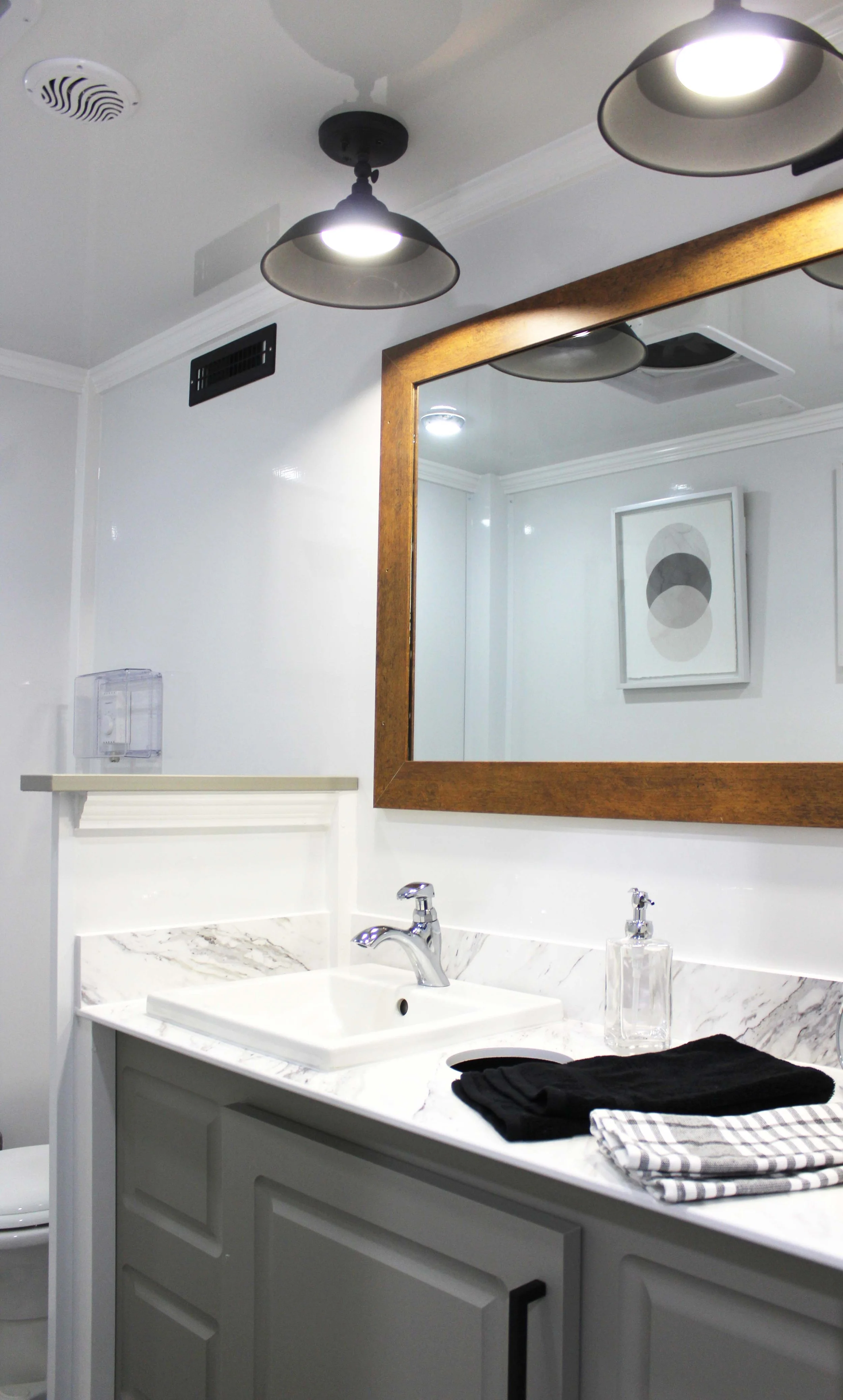 A bathroom with a white vanity, marble countertop, rectangular sink, and mirror with a wooden frame. There are black and checkered towels folded on the countertop. The wall has artwork with abstract circles. Overhead lighting fixtures are visible. A soap dispenser and a paper towel holder are also present.