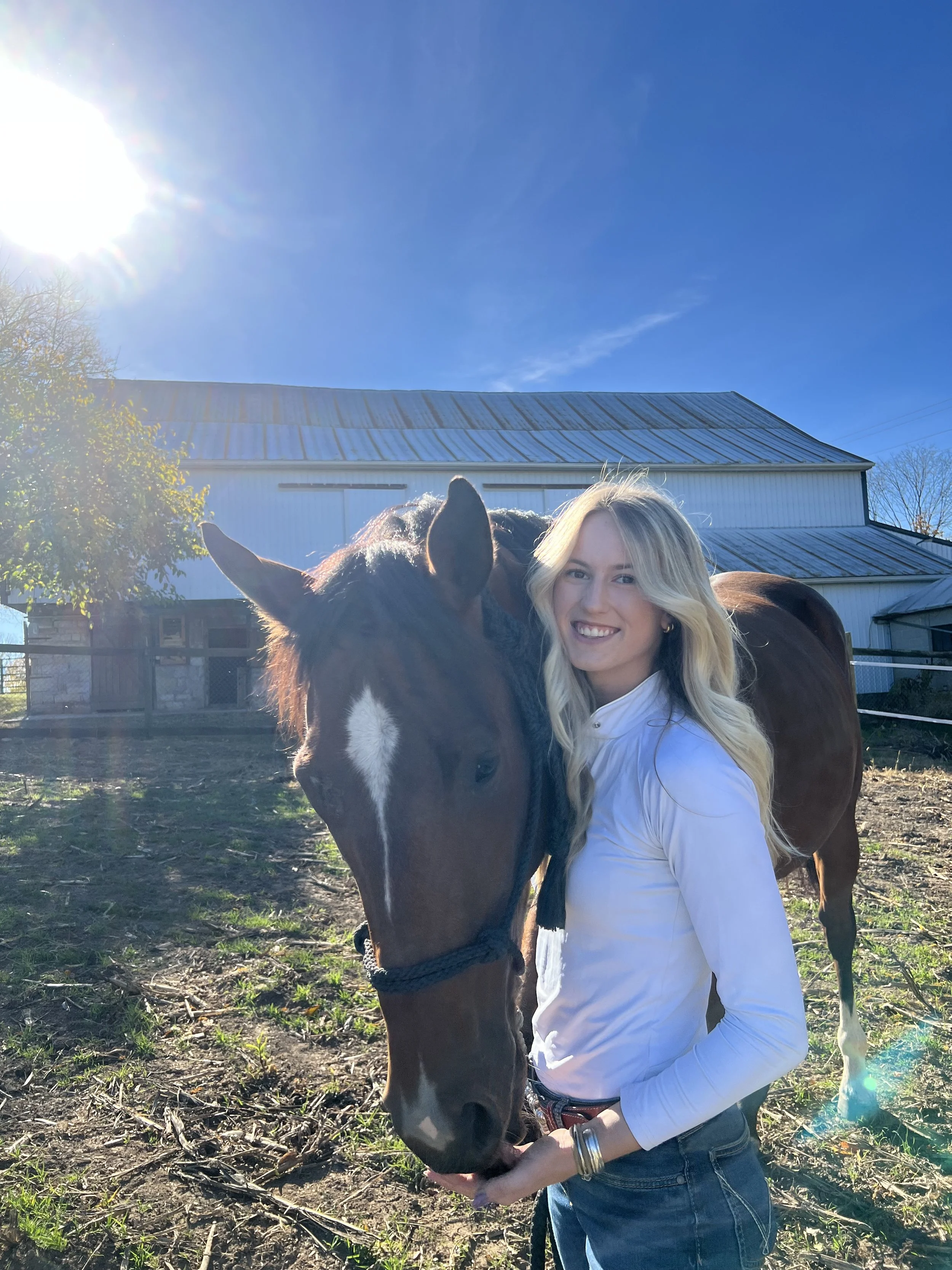 A woman with long blonde hair, wearing a white shirt, smiling and holding a brown horse's chin in an outdoor farm setting under a clear blue sky with sunlight.