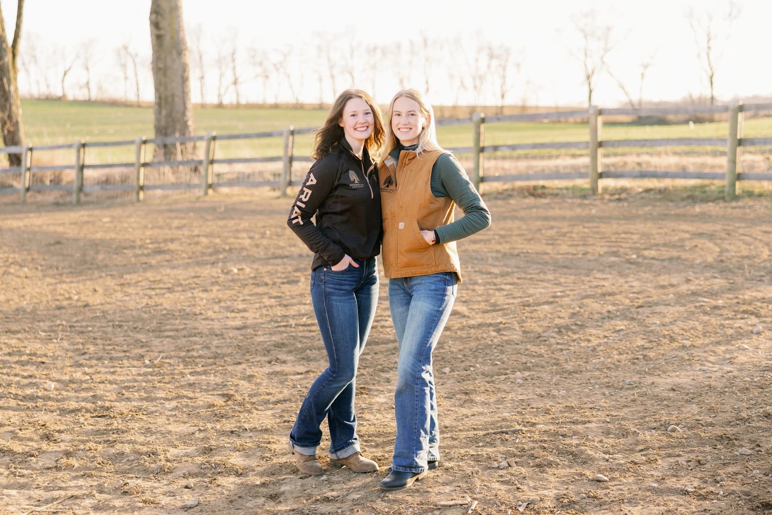 Two women stand close together in a farm field, smiling at the camera during sunset. One wears a black jacket and jeans, the other a tan vest and jeans. Wooden fence and leafless trees are in the background.