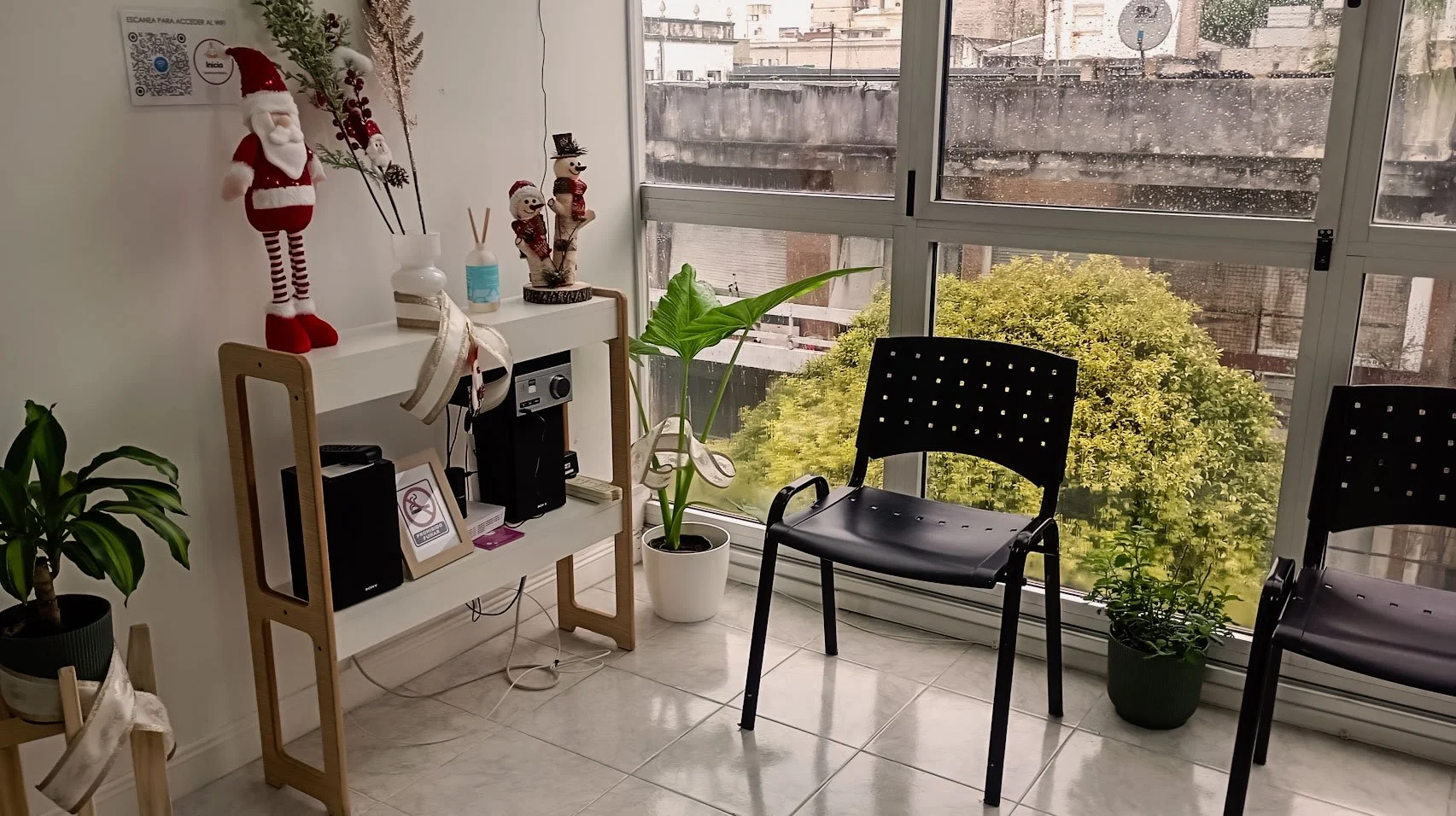 Un rincón luminoso de una oficina o sala de espera con decoración navideña, que incluye un muñeco de Papá Noel y figuras invernales, cerca de una ventana con vista a un paisaje urbano y un árbol verde, sillas negras, plantas en macetas