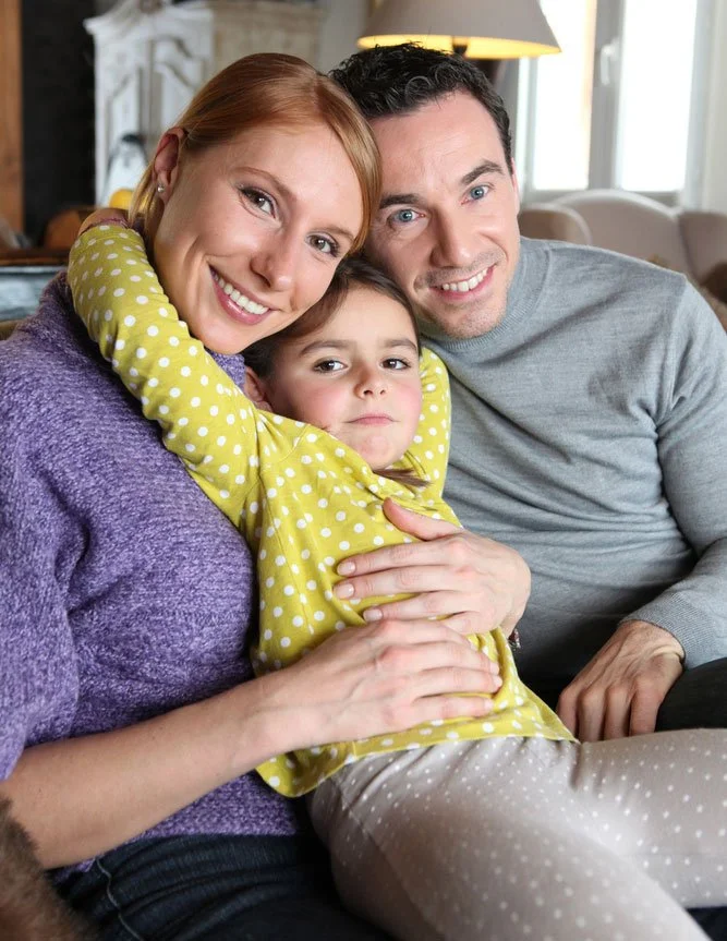 Una familia feliz de tres personas sentada en un sofá en una acogedora sala de estar. La madre, pelirroja, sonríe y viste un suéter morado. El padre, de cabello oscuro, sonríe y viste una camisa gris claro. Su hija pequeña, de cabello castaño.