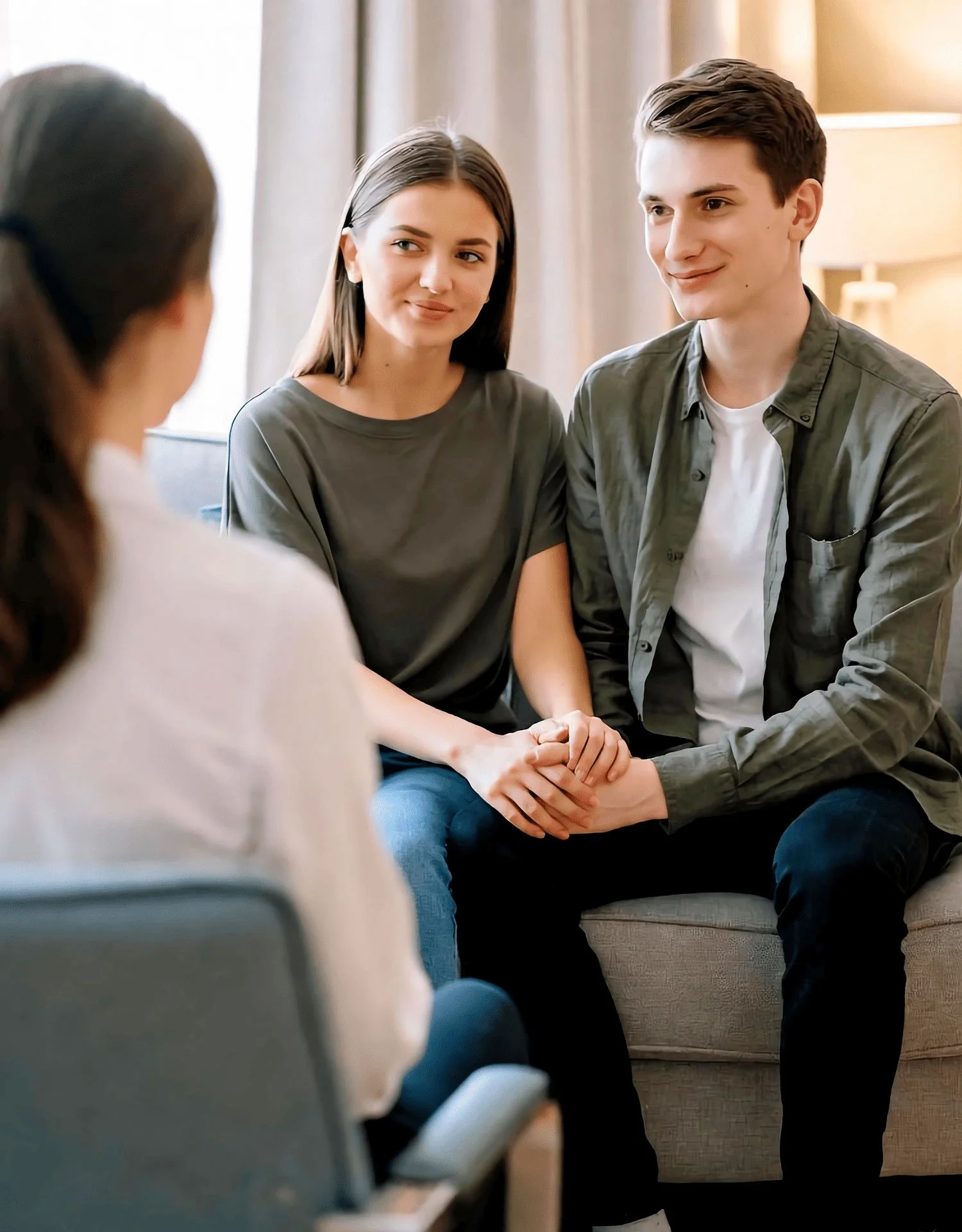 Una pareja joven sentada en un sofá, tomada de la mano, durante una sesión de asesoramiento con una terapeuta, vista desde atrás.