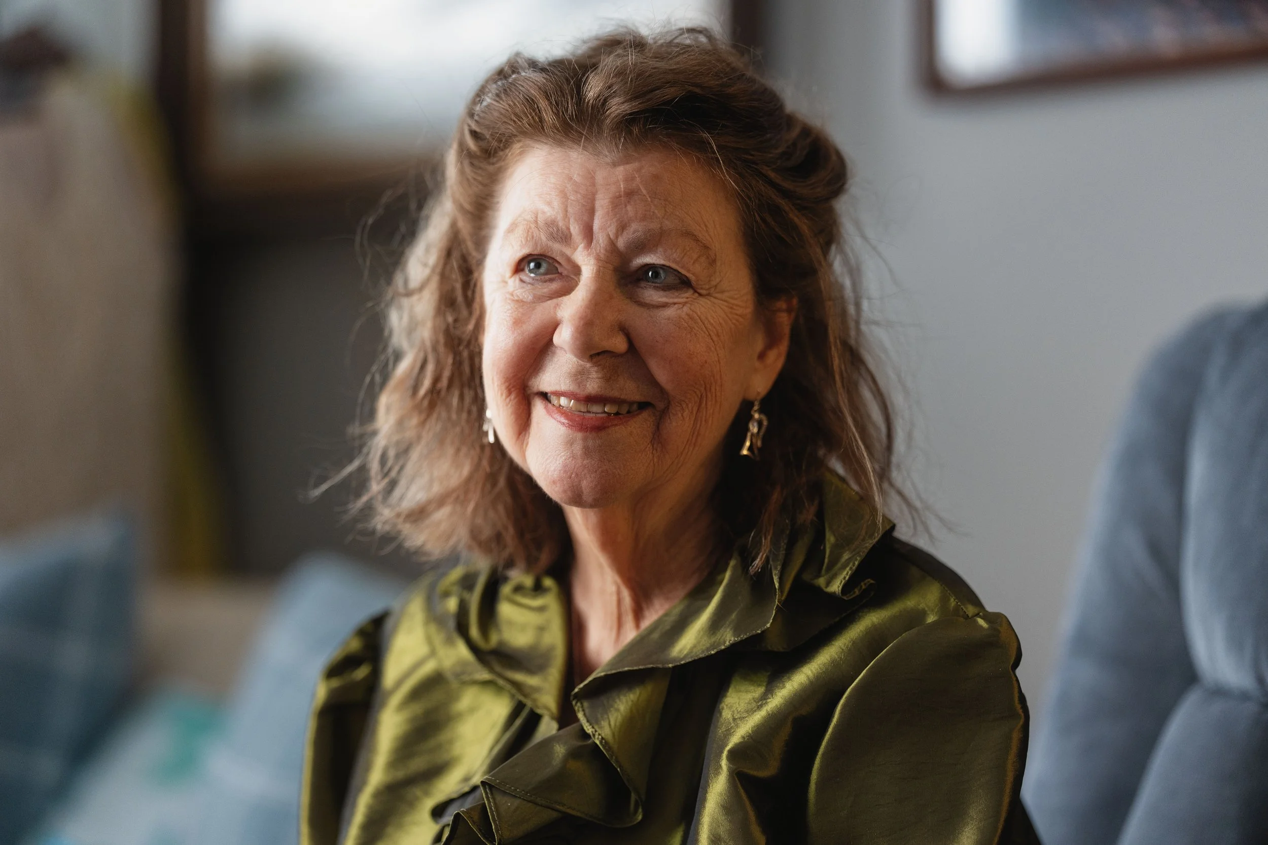 A smiling elderly woman with wavy brown hair and blue eyes, wearing gold earrings and a shiny olive green blouse, sitting indoors near a blue cushioned armchair.