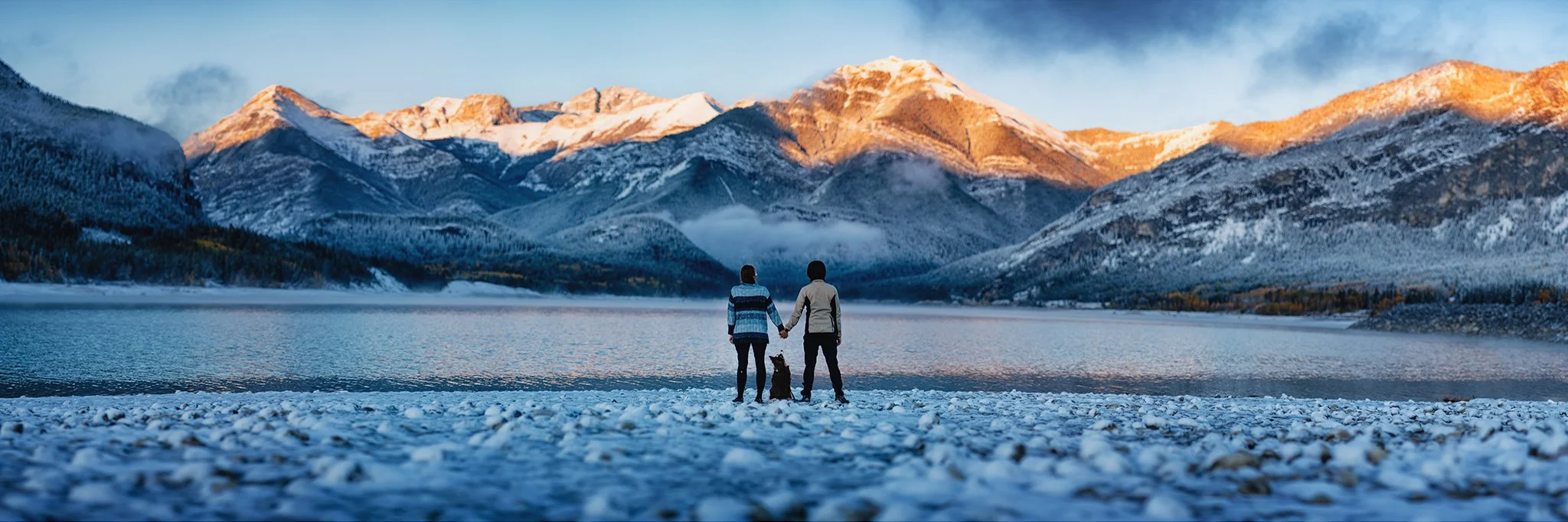 A couple and a dog standing on a snow-covered ground by a lake, holding hands, with snow-capped mountains in the background and a partly cloudy sky.