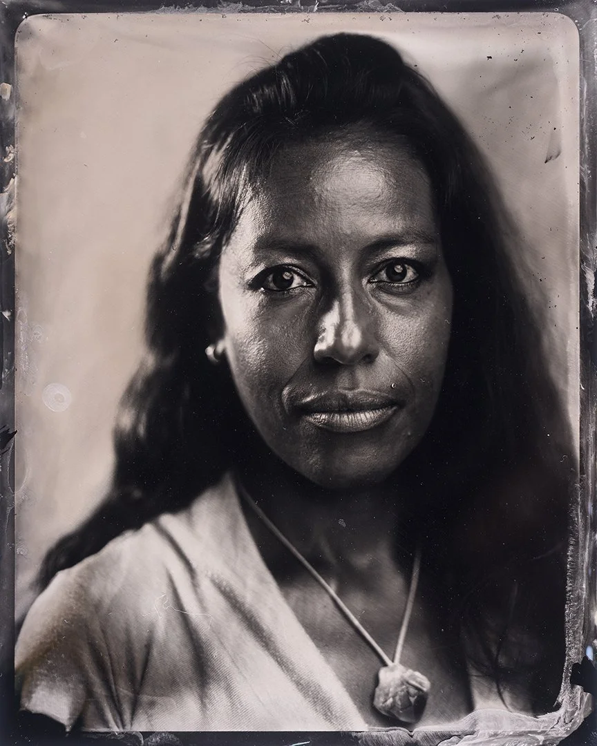 Black and white wet plate collodion tintype portrait photograph of a woman with long dark hair, wearing a necklace with a large pendant and a light-colored top.