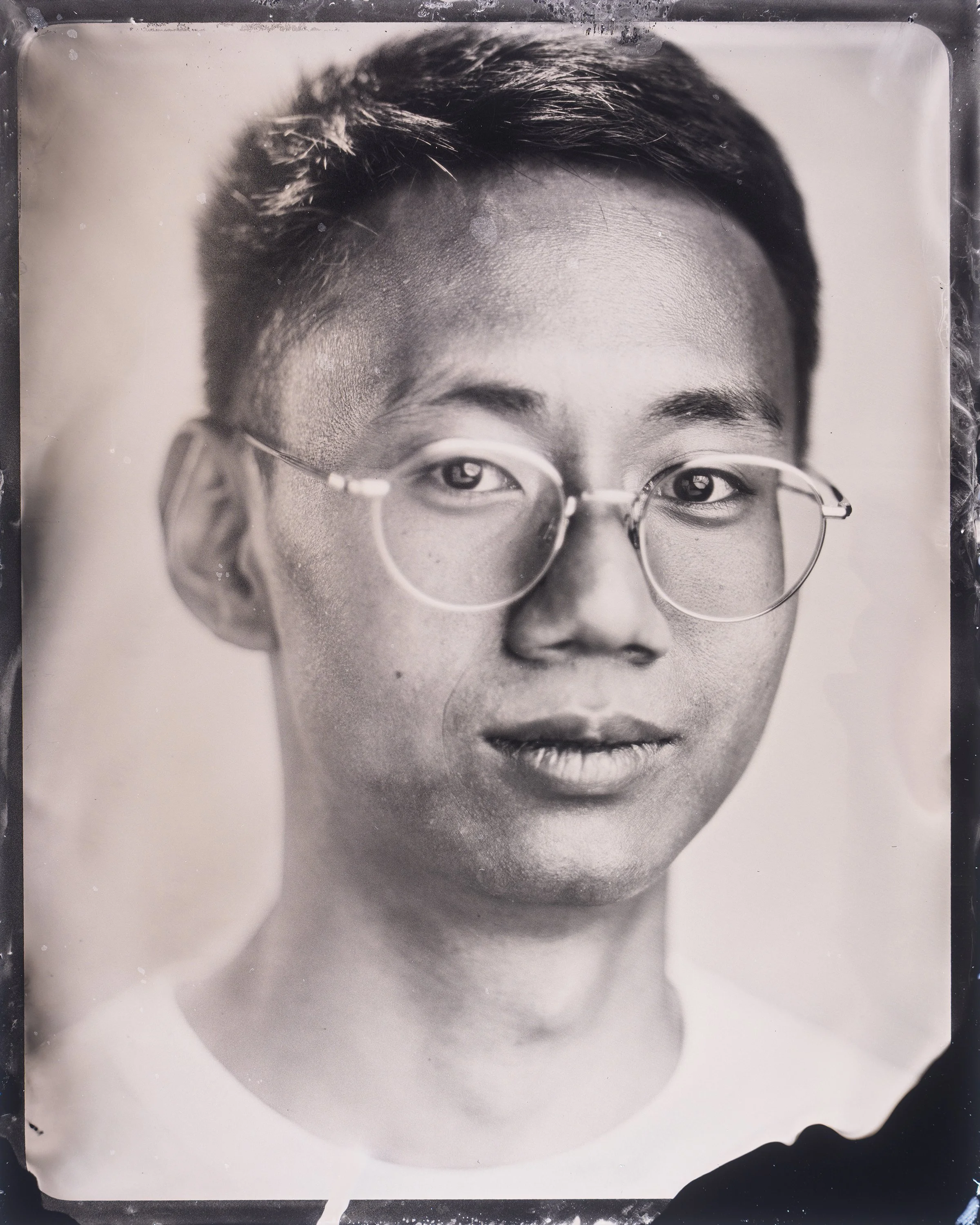 Black and white wet plate collodion tintype portrait photograph of a person's face wearing round glasses, with short hair and a neutral expression.