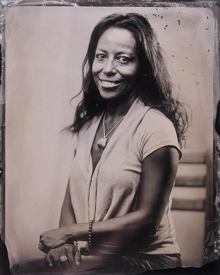 Black and white wet plate collodion tintype portrait of a smiling woman with wavy hair, wearing a short-sleeved shirt, necklace, and bracelet.