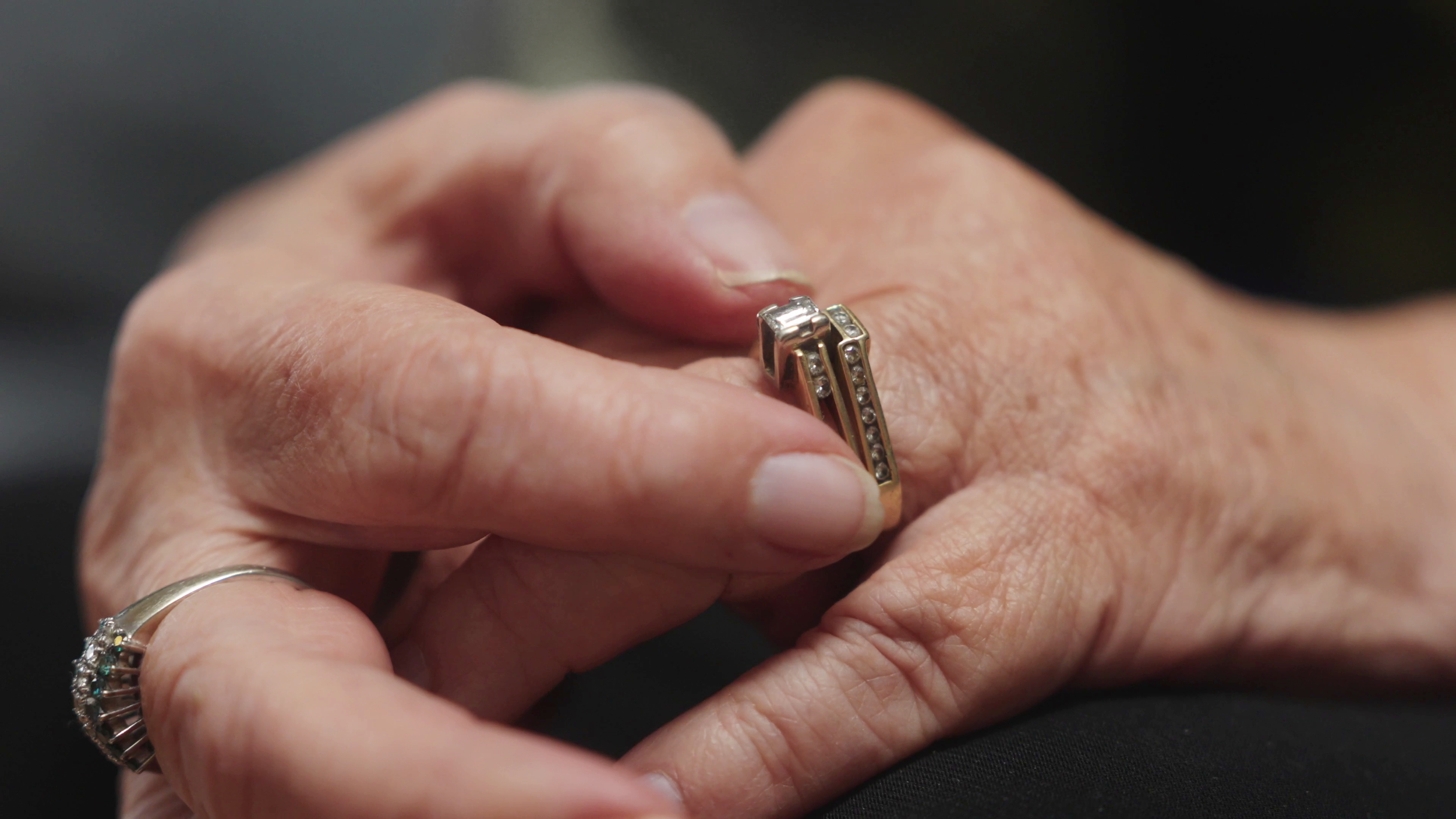 A pair of elderly hands with rings on fingers, one ring with diamonds and the other with multiple gemstones, holding a small, rectangular cut jewelry piece adorned with small diamonds.