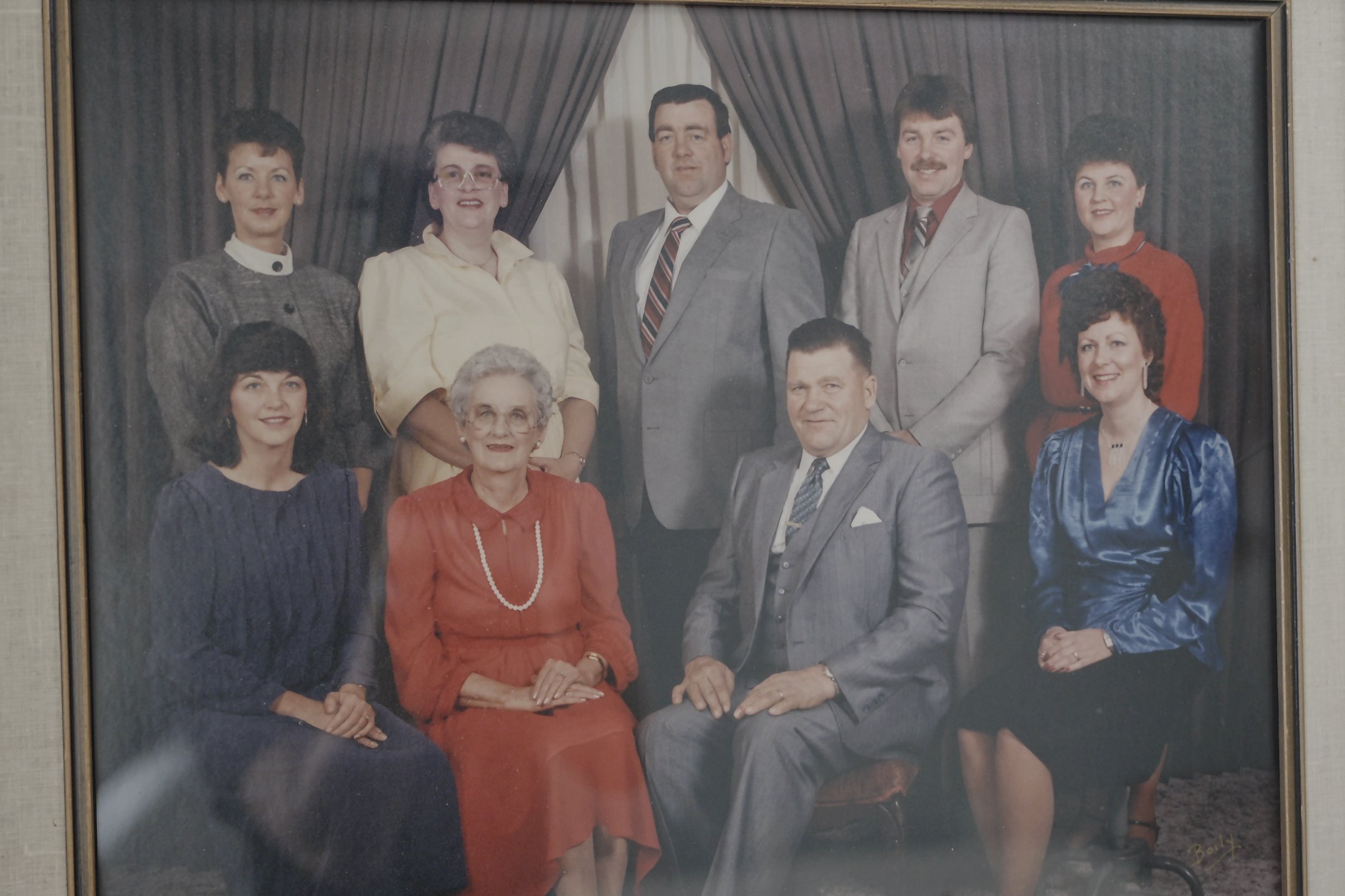 A vintage family portrait photograph featuring ten people, five women and five men, dressed in formal clothing. They are arranged with some standing and others seated, in front of a dark curtain backdrop.