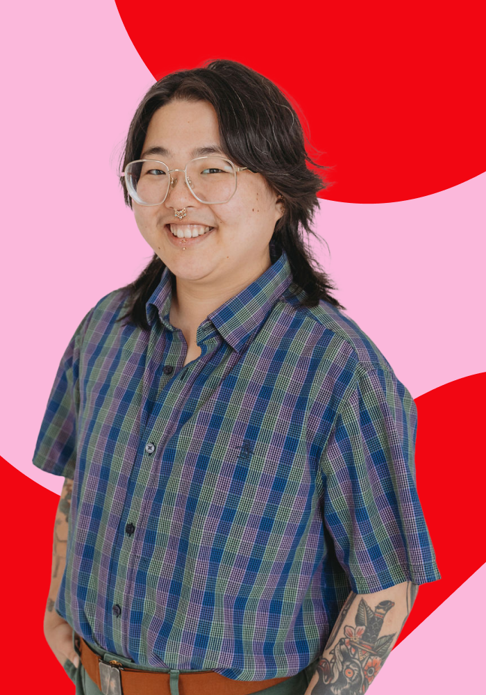 Portrait of a person with glasses and tattoos, smiling, wearing a colorful checkered shirt, standing against a vibrant background with pink and red abstract shapes.