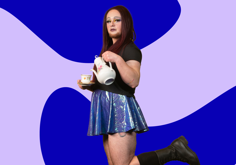 A person with long red hair and makeup, wearing a black shirt, shiny blue skirt, and black boots, holding a teapot and teacup, standing against a blue and purple abstract background.
