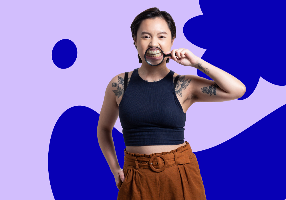 Young person with short dark hair, tattoos on arms, wearing a black tank top and brown high-waisted pants, looking through a magnifying glass with a playful expression, standing against a purple and dark blue abstract background.