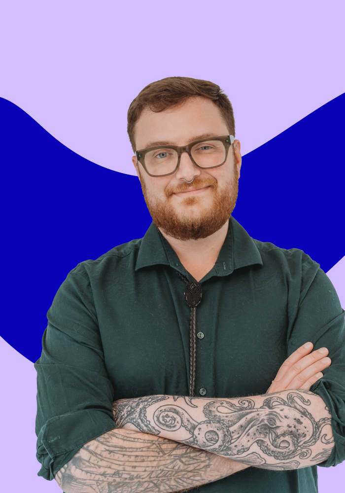 A young person with glasses, a beard, and tattoos on their arms crossed, wearing a dark green button-up shirt, standing against a purple background with blue abstract shapes.
