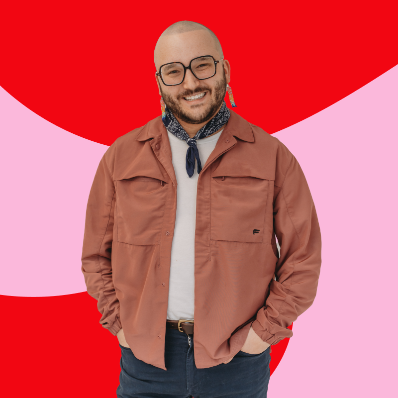 A smiling person with glasses and a beard, wearing a tan jacket, white shirt, and a patterned neckerchief, standing against a colorful background with red and pink shapes.
