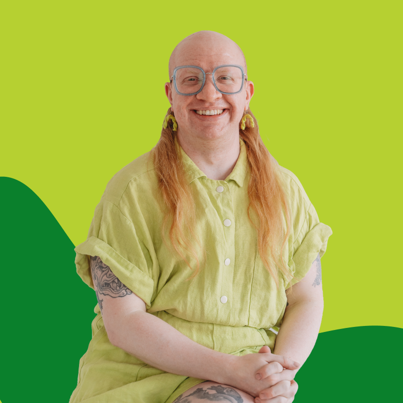 A smiling person with long red hair, wearing glasses and a light yellow shirt, posing against a vibrant green background.