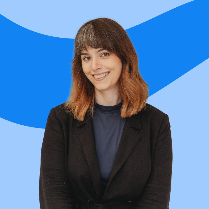 A person with shoulder-length brown hair and bangs, smiling, wearing a black blazer over a dark blue top, standing against a blue background with abstract shapes.