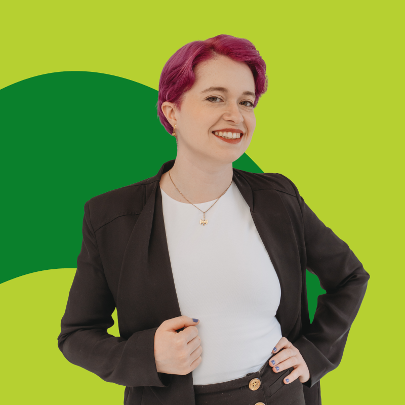 Person with short pink hair smiling, wearing a black blazer and white top against a green background.
