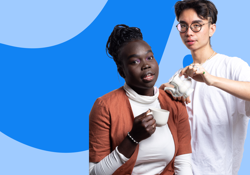 Two people standing close together against a blue geometric background. One person is holding a white teacup and looking at the camera, while the other person is pouring tea from a teapot.