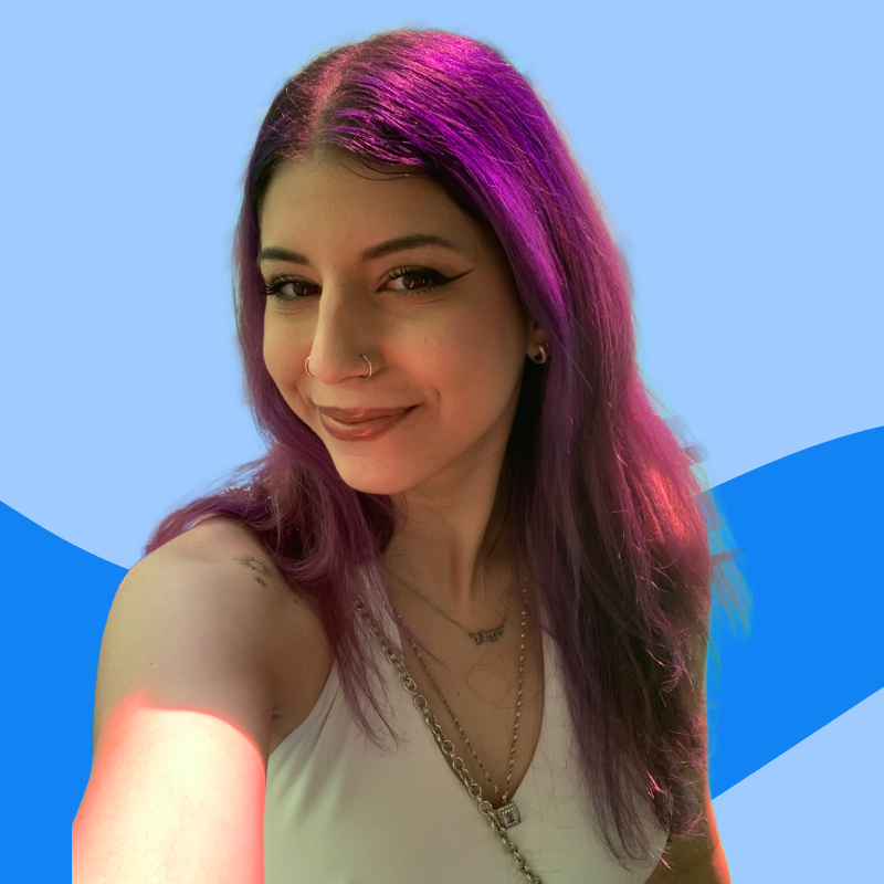 A person with purple and pink hair smiling at the camera, wearing earrings, necklaces, and a sleeveless top, with a light blue background.