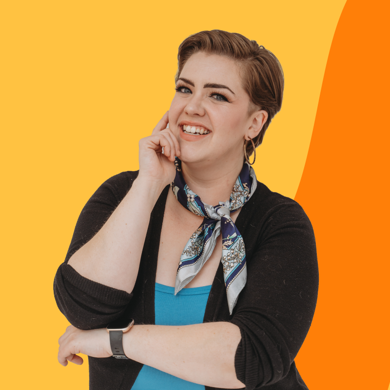 A smiling person with short hair, wearing a black cardigan, blue top, patterned scarf, and hoop earrings, posing with their hand on their face against an orange and yellow background.