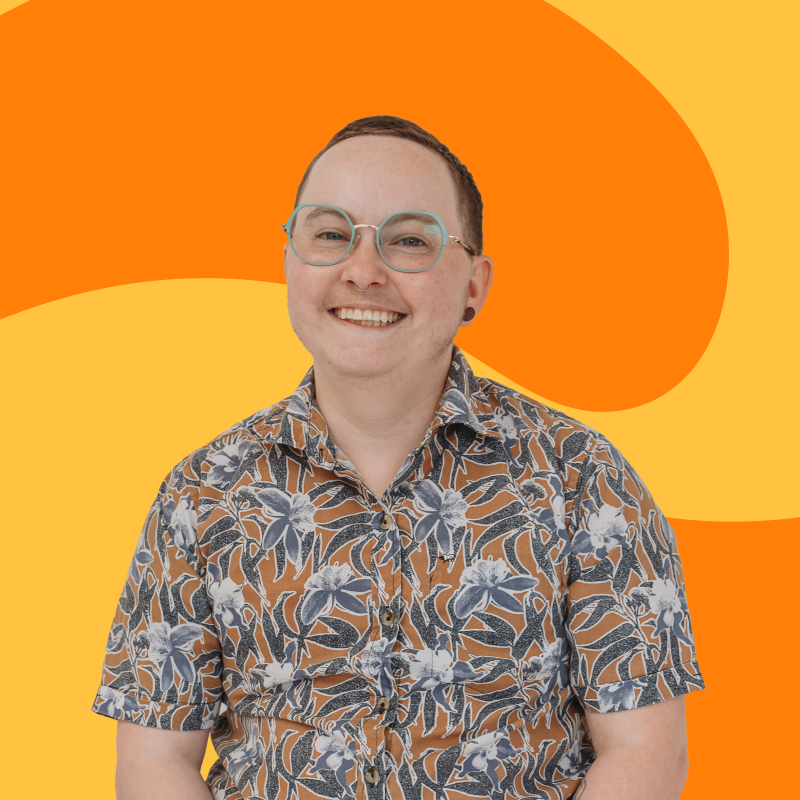 A person with short brown hair, glasses, and a septum piercing, smiling and wearing a short-sleeve button-up shirt with a floral pattern against a colorful, abstract background.