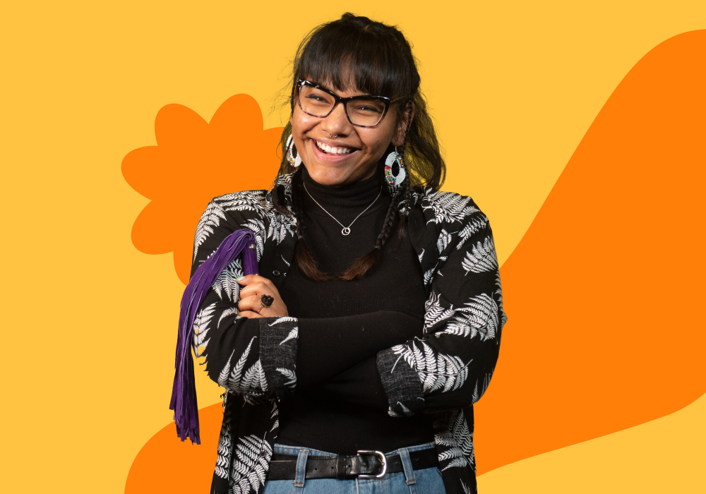 A young woman smiling, wearing glasses, a black turtleneck, and a black and white patterned jacket, standing with her arms crossed against a colorful abstract background.