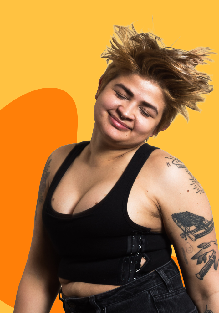 Young person with short, messy hair wearing a black crop top with suspenders and black jeans, smiling with eyes closed, standing against a yellow background with orange abstract shape.