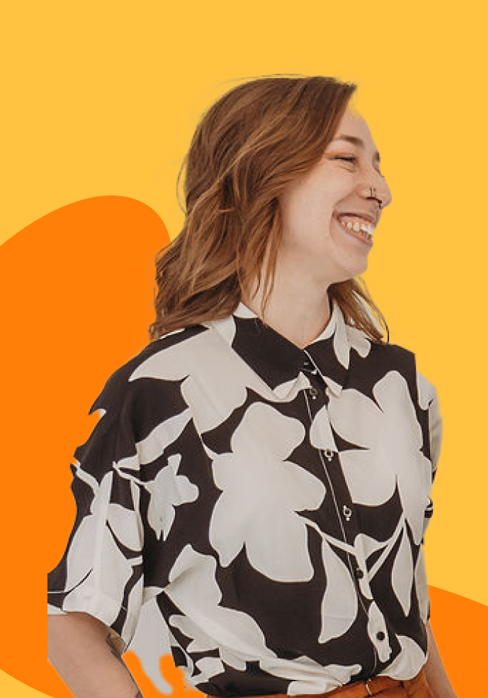 A young woman with red hair smiling and wearing a black and white floral patterned shirt, standing against a yellow and orange abstract background.