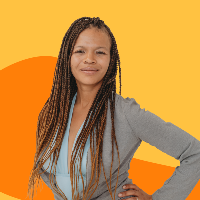 Person with long braided hair wearing a gray blazer and blue blouse posing confidently against a yellow and orange abstract background.