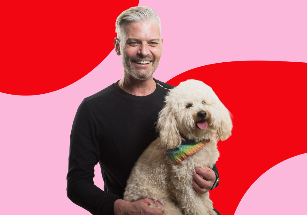 A smiling man with gray hair holding a fluffy white dog with a colorful bandana against a red and pink abstract background.