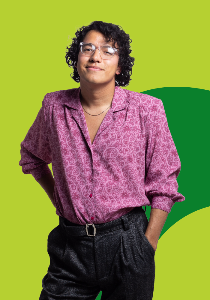 A young person with curly hair and clear glasses, wearing a pink patterned long-sleeve shirt tucked into black high-waisted trousers, standing with one hand in pocket against a bright green background.