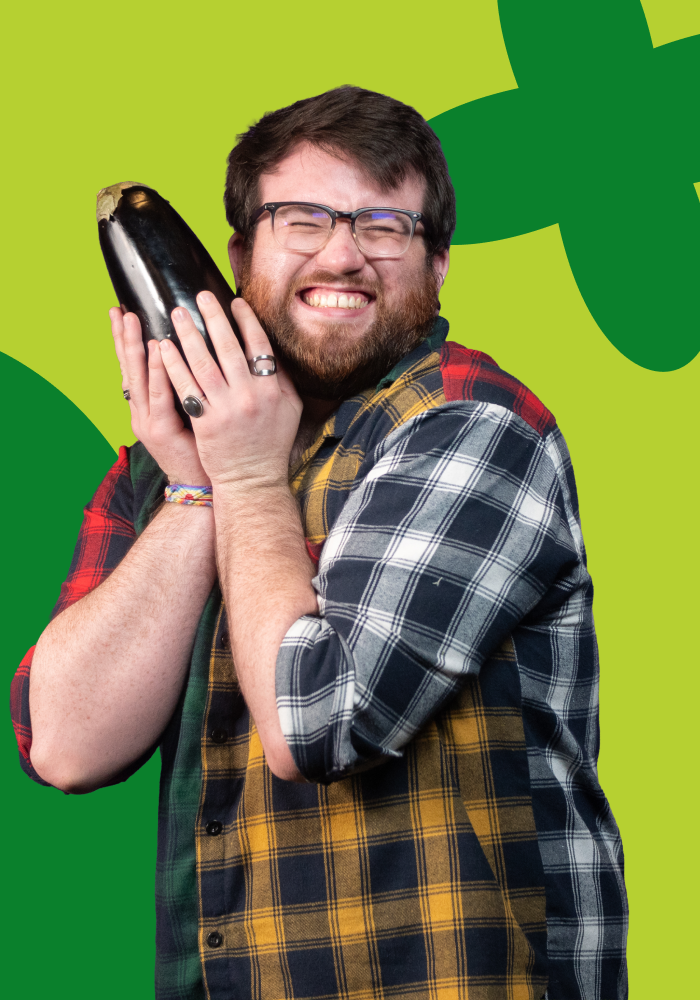 A smiling person with glasses and a beard holding a large eggplant close to their cheek, wearing a plaid shirt with a colorful background.