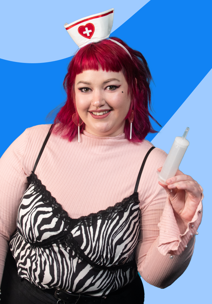 A woman with pink hair wearing a nurse hat, a pink ribbed shirt, and a zebra-print camisole, holding a nasal spray, smiling at the camera, in front of a blue background.