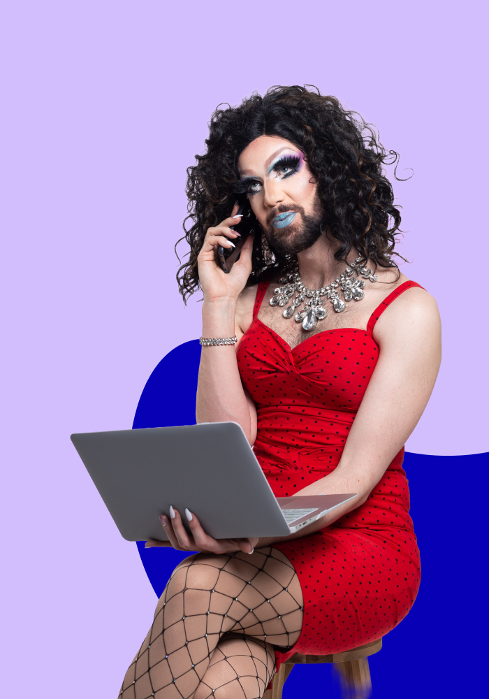 A person with curly dark hair, makeup, and blue lipstick, wearing a red polka dot dress, sitting on a stool with a laptop on their lap, talking on a mobile phone against a purple background.