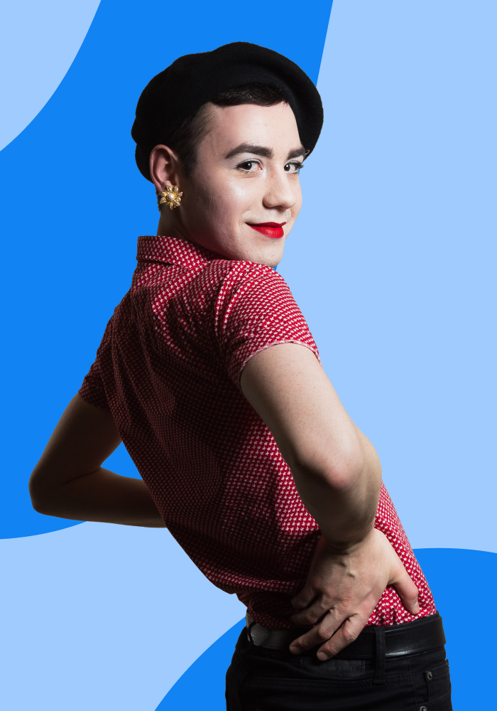 A person with short dark hair, wearing a black beret, gold floral earrings, red lipstick, a red polka dot shirt, standing against a vibrant blue and light blue background, smiling confidently.