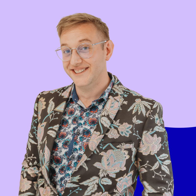 A smiling man with glasses wearing a floral patterned blazer and a colorful shirt against a pastel purple background.