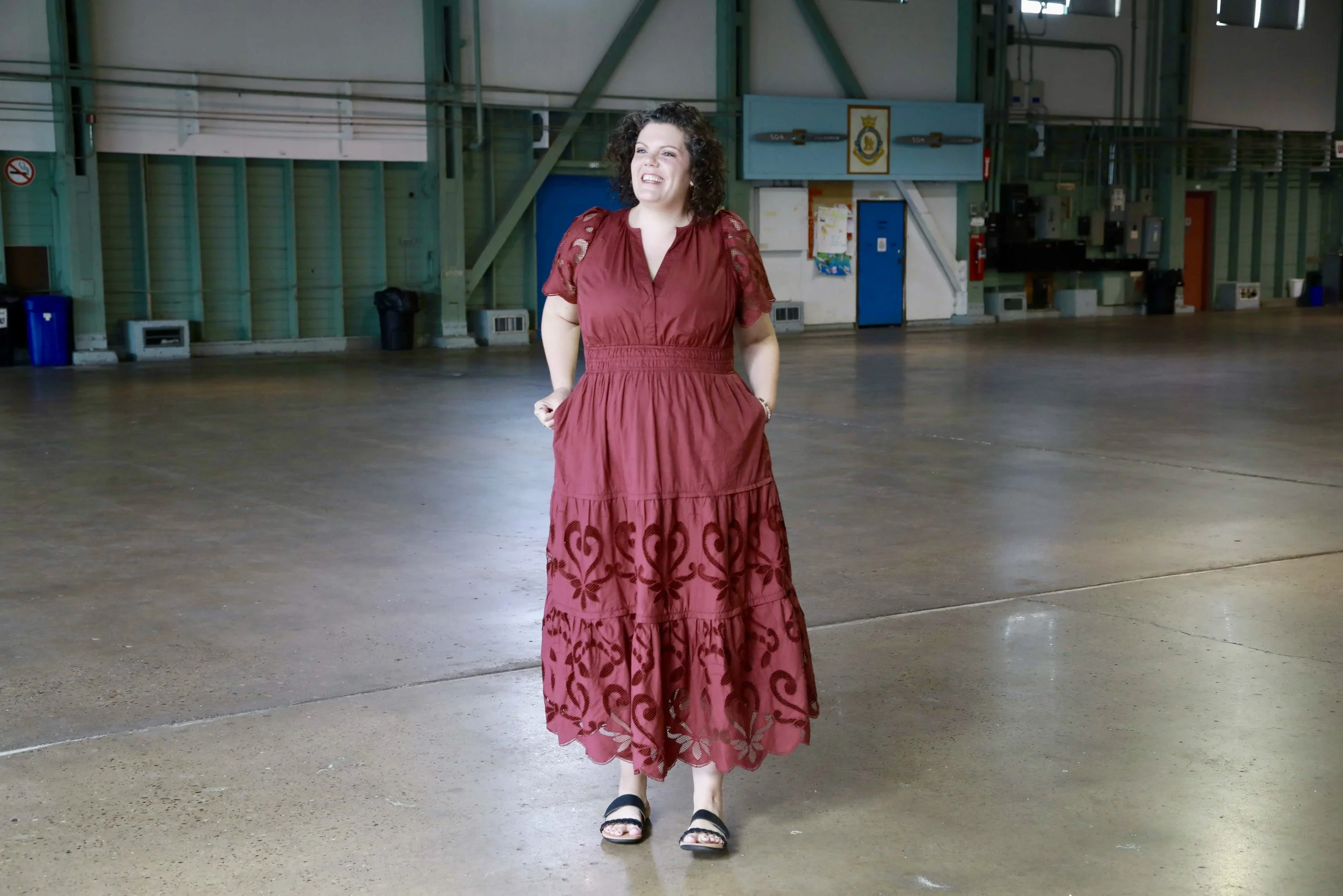 Author Catherine Cicuttini wearing long red dress standing in an empty indoor area with green and white walls, blue doors, and some machinery in the background.