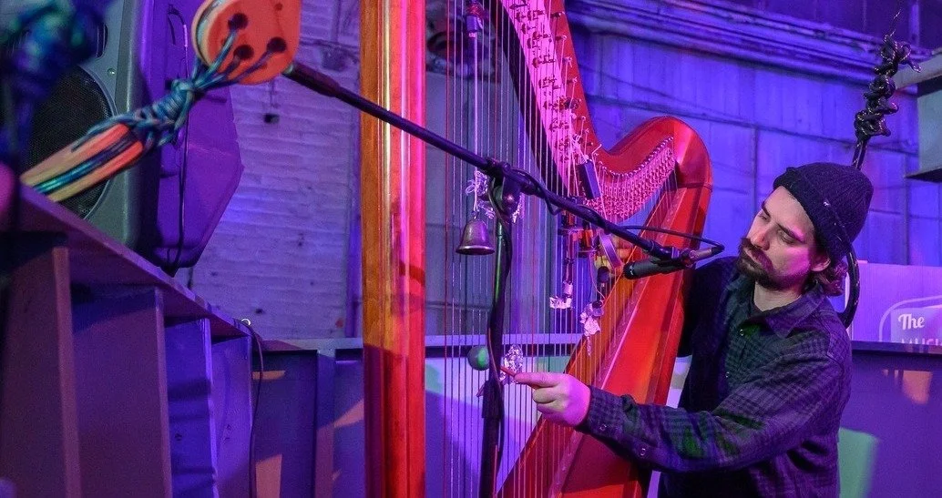 A man wearing a black beanie and dark shirt playing a large wooden harp in a colorful, neon-lit room.